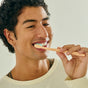 A smiling young man with curly hair brushing his teeth with a peach-colored toothbrush with blue bristles, wearing a cream waffle-knit shirt against a light background.