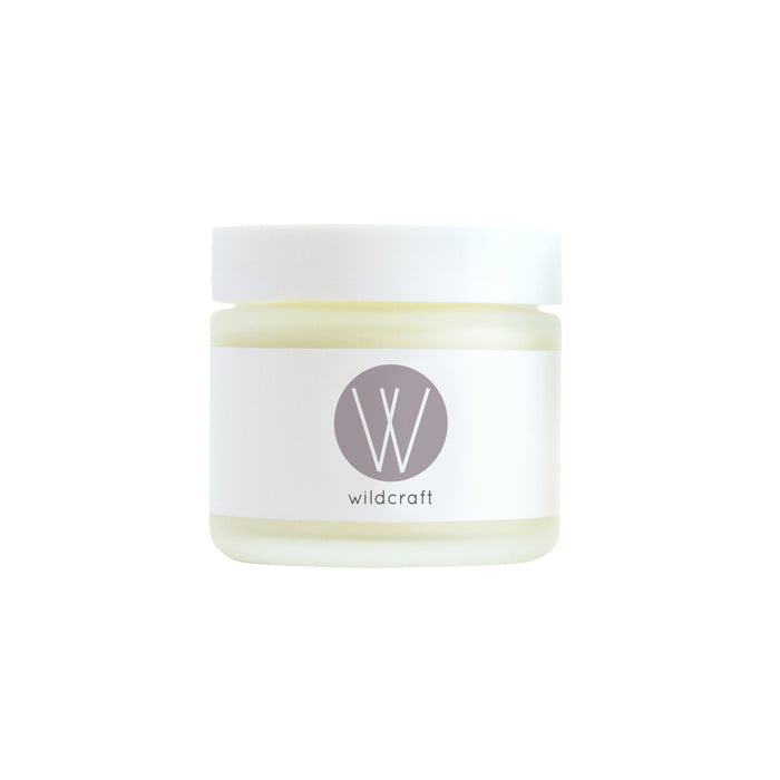 A small glass jar with a white lid and a minimalist label featuring a gray circle with a white W and the word wildcraft below it. The jar appears to contain a cream or balm.