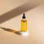 A clear glass dropper bottle labeled Terra, filled with yellow oil, stands on a white circular pedestal casting a shadow on a beige background with soft lighting.