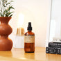 A brown glass spray bottle labeled Sandalwood Rose Room & Linen Spray sits on a wooden surface beside a terracotta vase with greenery, a glowing lamp, and a stack of books.