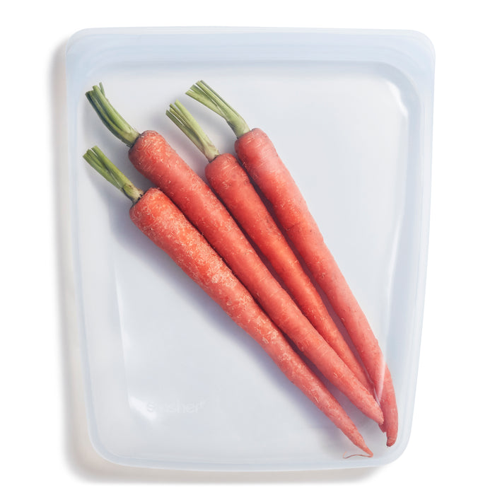 Four whole carrots with green stems are placed side by side inside a translucent, reusable silicone storage bag on a white background.