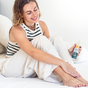 A woman in a striped tank top and light pants is sitting on a bed, applying Tree To Tubs Advanced Crack Defense Deep Moisturizing Heel & Foot Cream enriched with Urea to her leg. The bright, airy setting offers the perfect atmosphere for caring for dry, cracked heels.