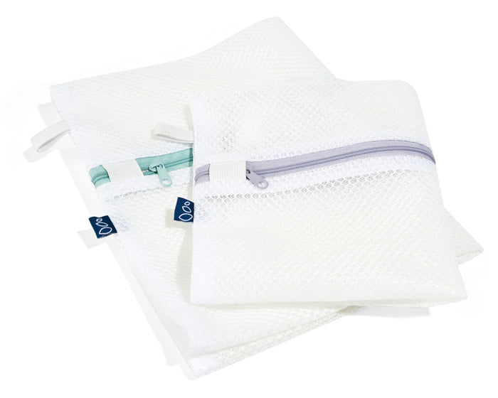 Two white mesh zippered bags with colored zippers—one with a teal zipper, one with a light gray zipper—overlapping each other. Each bag has a small blue fabric tag on the side.