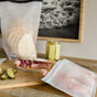 A ham and red onion sandwich rests on a wooden board next to pickle slices and a (re)zip Reusable Bread, Bagel & Pantry Roll Top Bag. A jar of pickles and a framed photo appear in the background.