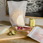 A ham, cheese, and pickle sandwich rests on a wooden board beside a (re)zip Reusable Bread, Bagel & Pantry Roll Top Bag, cheese slices, deli meat, and pickles. A black-and-white photo hangs on the wall in the background.