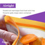 A hand seals an orange (re)zip Reusable Stand-Up Bag filled with sliced carrots. Text reads: Airtight double lock seal keeps food fresh and crisp. Made from food-grade PEVA for leakproof, odor-controlling storage. (re)zip logo in the corner.
