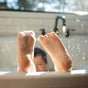 A child is taking a bubble bath, with their feet covered in soap suds and raised over the edge of the tub. Sunlight streams in, creating a bright and playful atmosphere. The childs face is partially visible in the background.