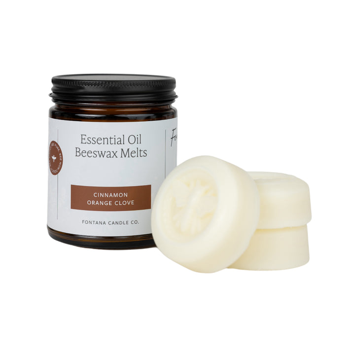 A brown jar labeled “Essential Oil Beeswax Melts—Cinnamon Orange Clove” sits next to three round, off-white wax melts stacked on one another. The melts have a bee design engraved on the top.