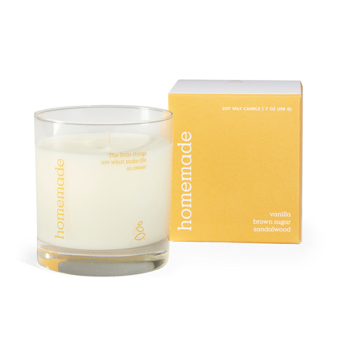 A white soy wax candle in a clear glass jar sits beside a yellow box labeled homemade. The box lists the scent as vanilla, brown sugar, sandalwood. The candle jar has a small quote printed in yellow.