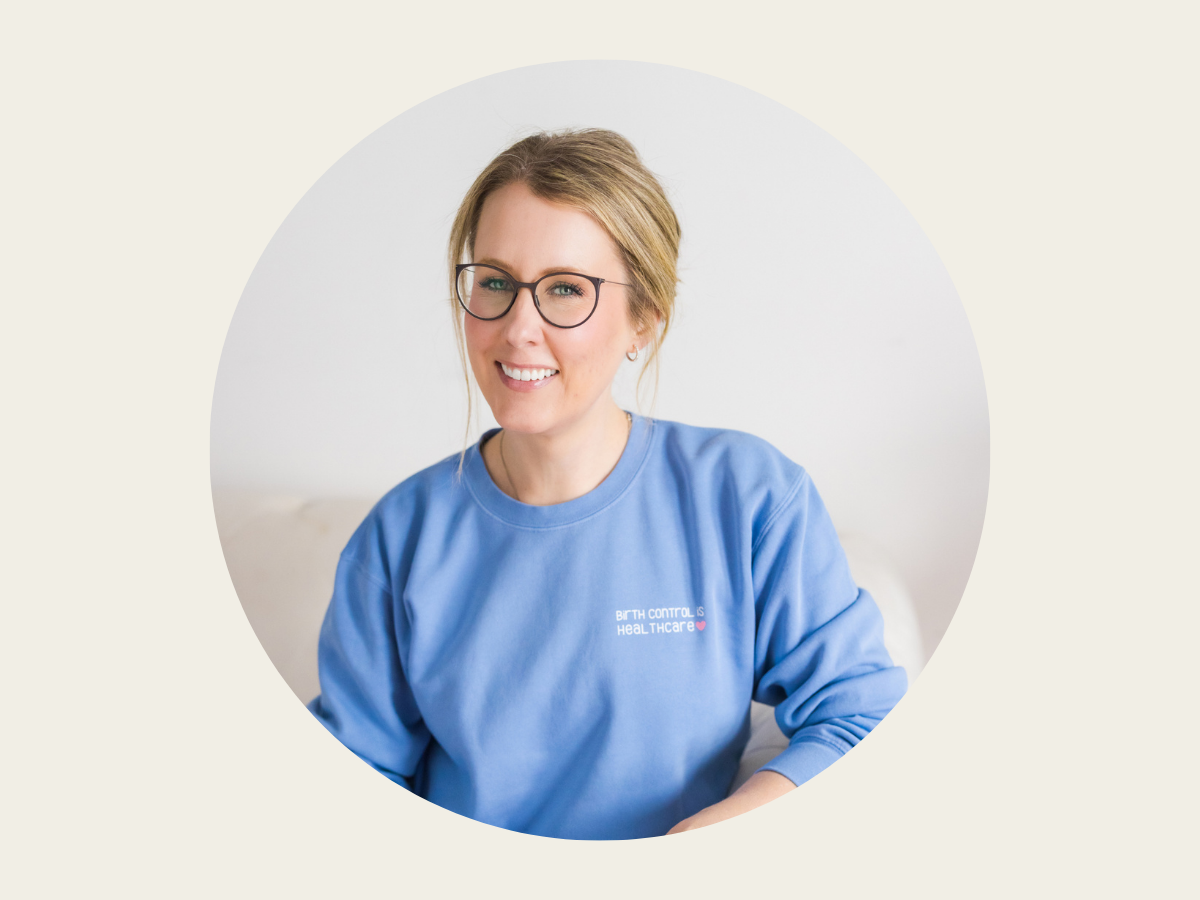 A woman with long blonde hair in a ponytail, wearing glasses and a light blue sweatshirt, smiles while sitting on a light-colored couch against a plain background.