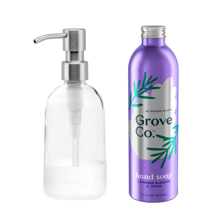 A clear glass hand soap dispenser with a silver pump sits beside a purple metal Grove Co. Hand Soap Starter Set bottle, featuring artful green leaf designs and scented with lavender blossom and thyme, perfect for refills.