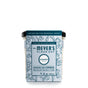 A Mrs. Meyers Soy Tin Candle, infused with essential oils, features a blue label with snowdrop flowers reading Snowdrop Scent and Limited Edition Seasonal Scent. It has a 35-hour burn time and is housed in a transparent square jar with a black lid.