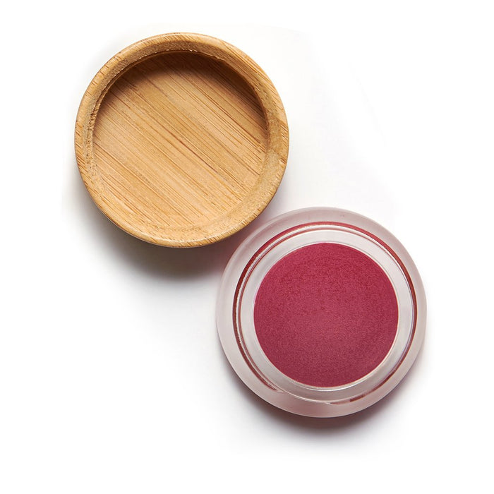 A small round glass container with a deep pink cream inside, placed next to a flat, circular wooden lid on a white background.