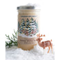 A jar of Savannah Bee Company Winter White Whipped Honey with a creamy texture rests on snow, next to greenery and a toy deer. The illustrated label features snowy trees and red berries, while snow covers the lid and ground.