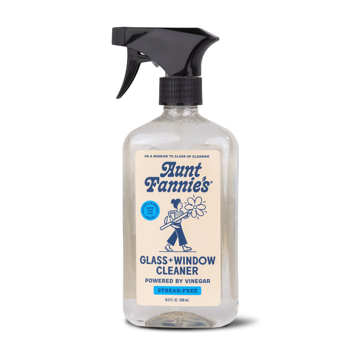 A clear spray bottle of Aunt Fannie’s Glass + Window Cleaner with a black nozzle. The label features a blue illustration of a woman with flowers and text stating its streak-free and powered by vinegar.