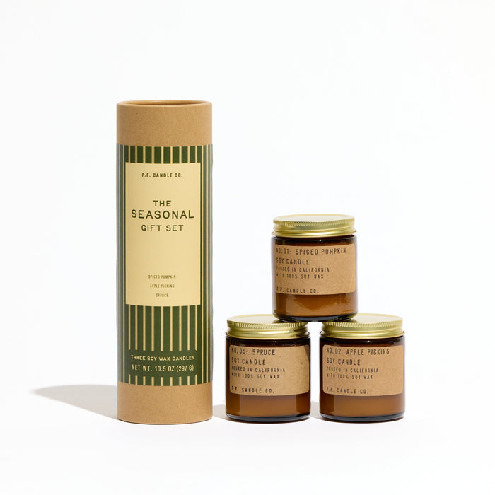 A Seasonal Mini Candle Gift Set featuring a cylindrical cardboard tube next to three mini candles in amber glass with gold lids.