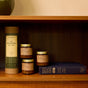 Three small amber glass mini candles and a cylindrical gift tube from the P.F. Candle Co. Seasonal Mini Candle Gift Set sitting on a wooden bookshelf next to a hardcover book.
