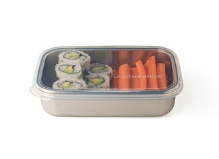 A stainless steel lunch container with a clear lid contains four avocado sushi rolls on the left and several carrot sticks on the right.