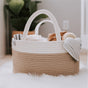A woven basket with white and tan stripes sits on a fluffy white rug, holding folded baby clothes and a gray blanket, with a leafy plant and wall outlet in the background.