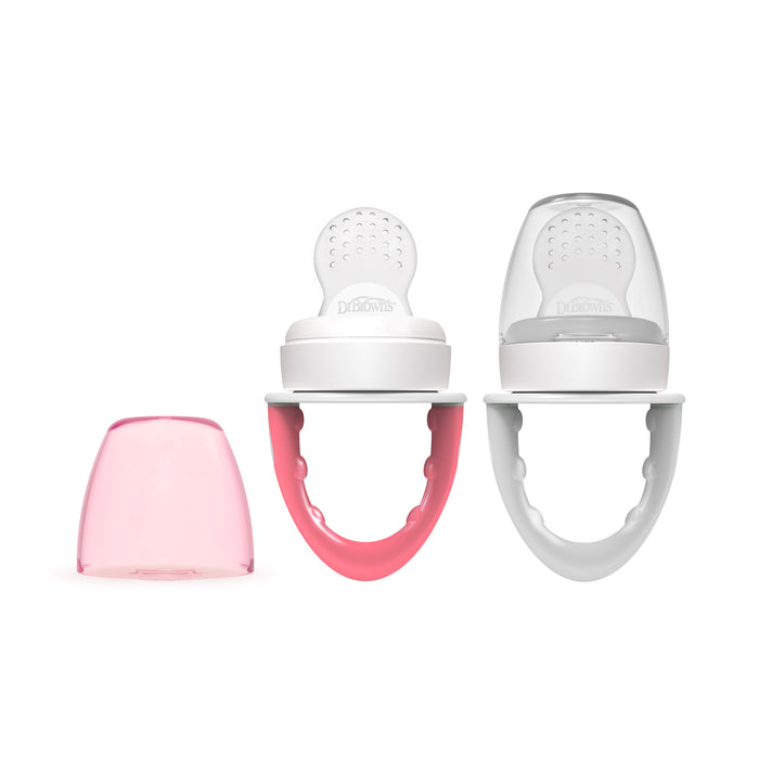 Two baby fruit feeders with rounded handles, one with a pink handle and the other white, are shown standing upright. A translucent pink cap is placed next to them on a white background.