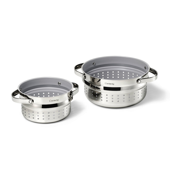 Two stainless steel colanders with gray interiors and side handles, one smaller and one larger, both featuring round holes for draining. The brand name Caraway is visible on the front of each colander.