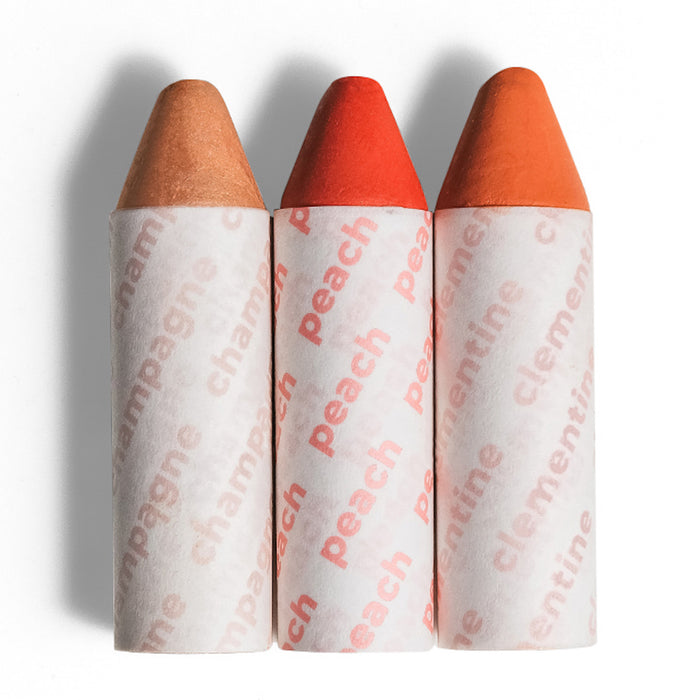 Three crayon-shaped lip balms stand upright side by side, wrapped in white paper labeled “champagne,” “peach,” and “clementine.” The colors range from nude to bright orange and coral.