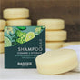 A box of Badger Balm All-In-One Shampoo & Conditioner Bar - Cleanse & Hydrate sits next to an oval organic shampoo bar, with other bars stacked in the background. The packaging features green leaves, citrus fruits, and product details.
