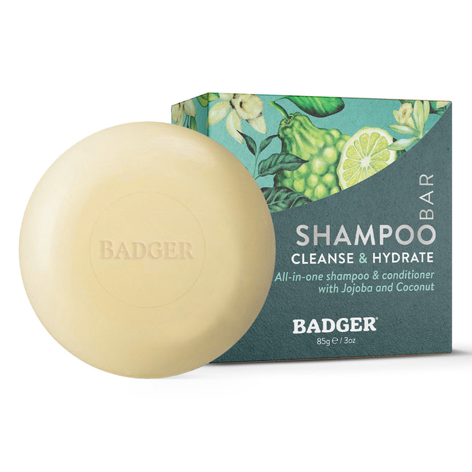 A round beige All-In-One Shampoo & Conditioner Bar - Cleanse & Hydrate from Badger Balm, featuring a BADGER emboss and crafted with natural ingredients, is pictured beside its green-and-blue box adorned with botanical illustrations and white text displaying product details and the SHAMPOO BAR: Cleanse & Hydrate label.