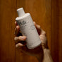 A hand holds a white bottle of Phil's Strengthening + Thickening Shampoo—ideal for scalp health and thick hair—covered in water droplets, set against a wooden background.