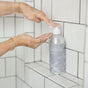 A person pumps Plaine Products Shampoo, Unscented from a silver bottle labeled "Refill • Reuse • Replace Shampoo" onto their hand in a tiled shower.