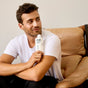 A man with short brown hair and a beard, wearing a white T-shirt, sits on a tan leather chair holding a bottle of Phil's Strengthening + Thickening Shampoo near his face, smiling slightly as he cares for scalp health and thicker hair.