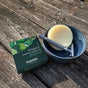 The Badger Balm Shave Bar - Smooth & Moisturize sits in a blue bowl alongside a razor, with its green Badger Balm packaging resting on a weathered wooden surface. Sunlight casts shadows across the plastic-free scene.