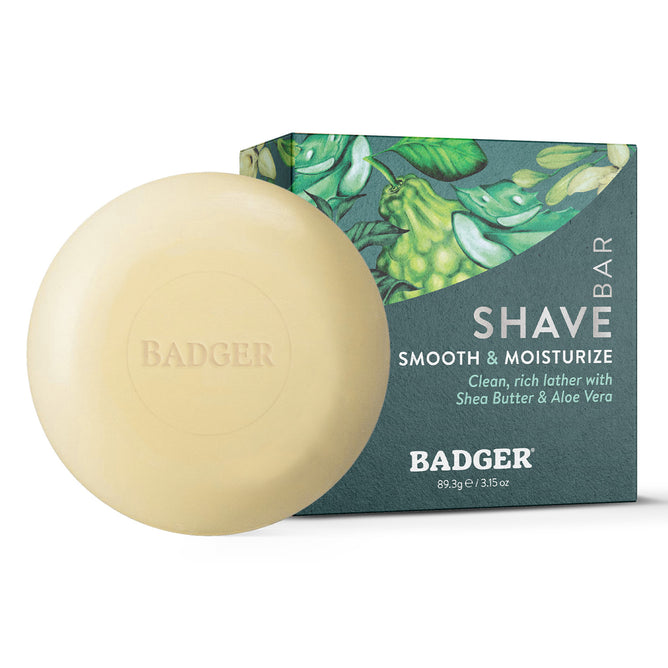 A round Badger Balm Shave Bar - Smooth & Moisturize sits in front of its packaging, which displays green botanical artwork and product text highlighting shea butter and aloe vera for smooth, moisturized, plastic-free shaving.