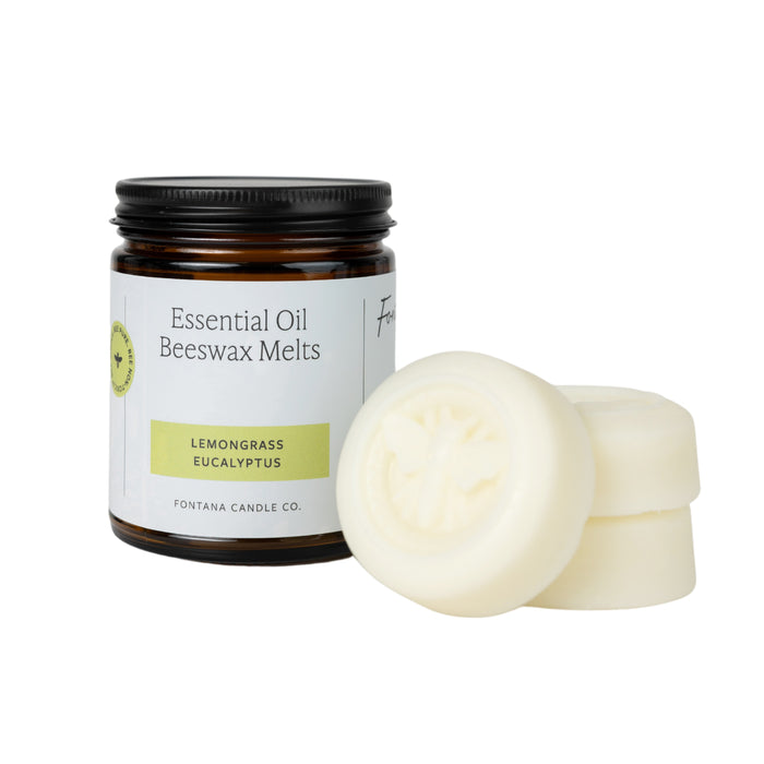A brown glass jar labeled Essential Oil Beeswax Melts Lemongrass Eucalyptus by Fontana Candle Co. sits next to three white beeswax melts, one of which features a bee design on top.