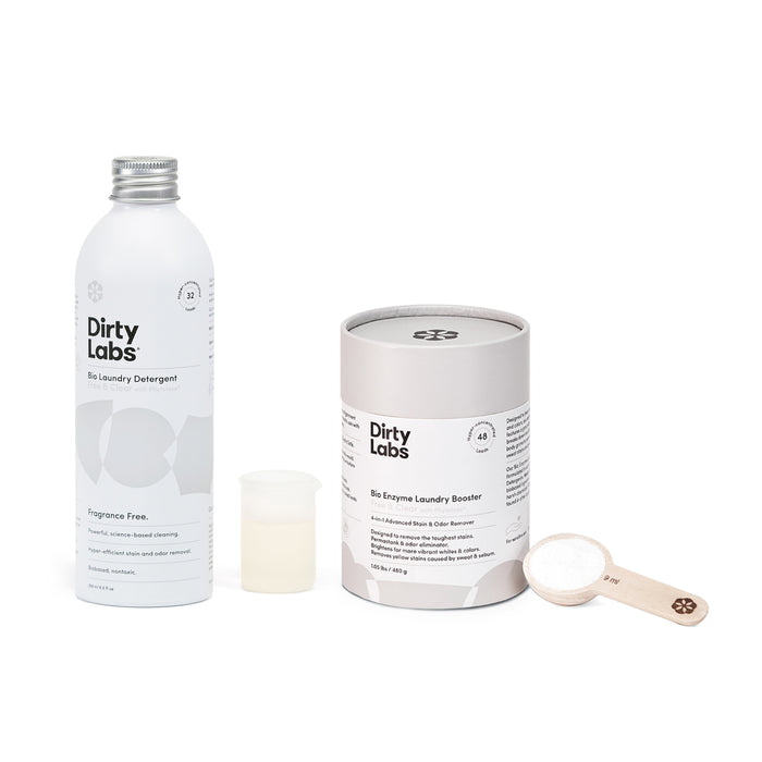 A Dirty Labs scene features the Bio Laundry Detergent with Phytolase® in a white bottle with a silver cap beside a cylindrical Bio Enzyme Laundry Booster, highlighting clean science. A small measuring cup and wooden scoop with enzyme-rich powder complete the set.