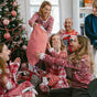 A family in matching festive pajamas exchanges gifts by a decorated Christmas tree. Three smiling girls sit excitedly as a woman hands them a large striped present, while two men watch and smile in the background.