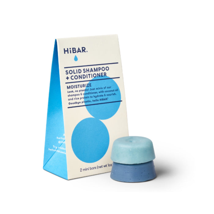A blue and cream triangular HiBAR box labeled Solid Shampoo + Conditioner Moisturize stands next to a round, two-tone blue shampoo and conditioner bar. The packaging highlights its plastic-free and natural qualities.