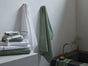 A bathroom featuring a stack of Coyuchi Air Weight® Organic Towels in green and white neatly arranged on the counter, two highly absorbent bath towels from the collection hanging on the wall, an organic cotton towel by Coyuchi draped over the edge of the bathtub, and a small vase with flowers placed near the faucet.