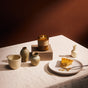 A Seasonal Mini Candle Gift Set by P.F. Candle Co. in an amber glass jar sits on a stone block next to a slice of pumpkin pie on a plate, surrounded by small ceramic vases on a tablecloth.