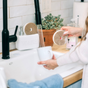 Someone washes a white mug in the kitchen sink with a Scrub Be Love Round Heart Dish Sponge (4-Pack) under running water, with soap, a plant, and other kitchen items on the countertop.