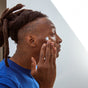 A person with dreadlocks and a tattoo on the side of their head applies face cream to their cheek while wearing a blue shirt, seen in profile against a light background.