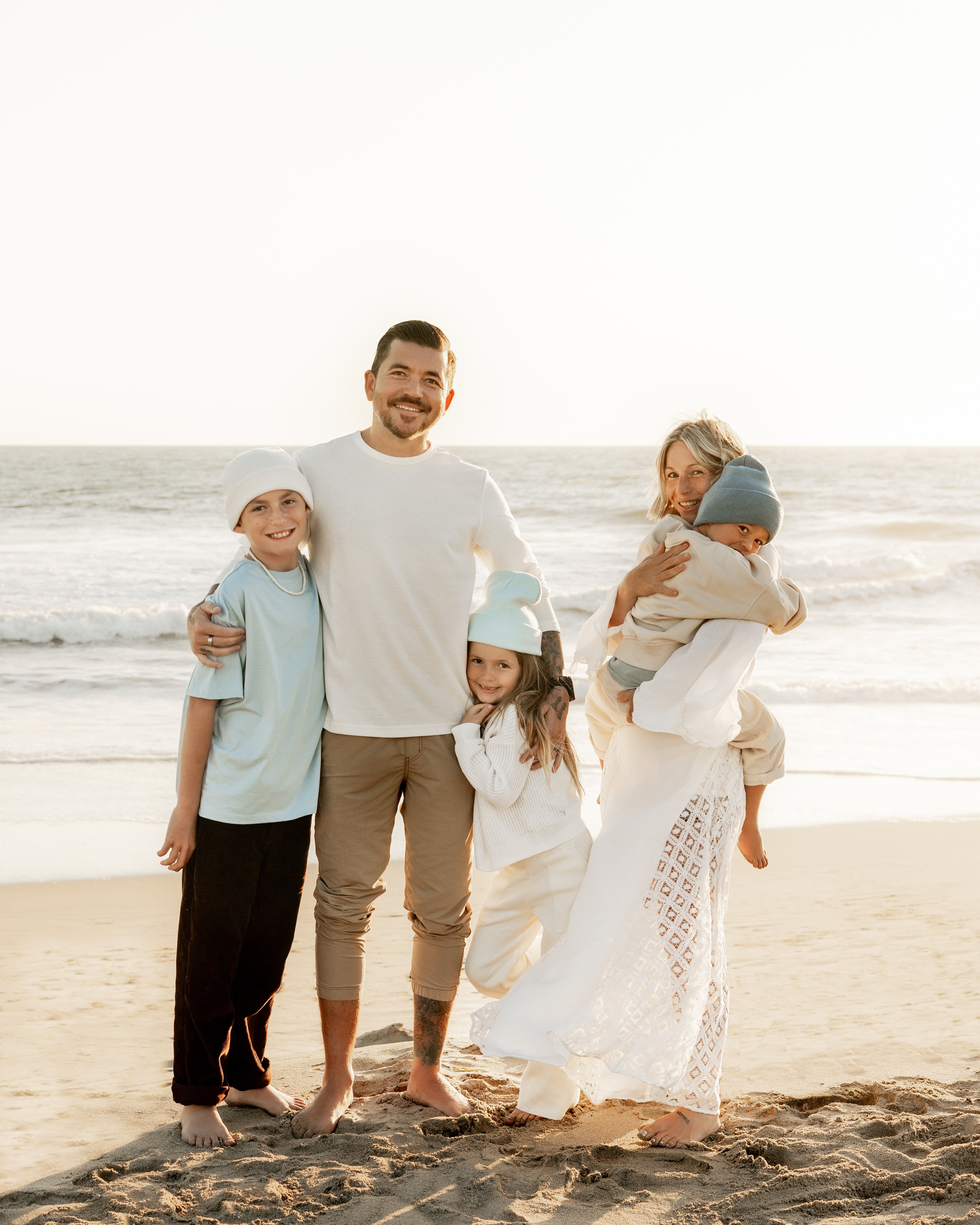 A smiling family of four stands on a sandy beach at sunset. The father hugs an older boy and a young girl, while the mother holds a small child. The ocean waves and sky are visible in the background.