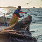 A fisherman in yellow overalls hauls a net of responsibly sourced White Anchovies Spicy for Patagonia Provisions on a boat at sea, as birds soar overhead and the sun sets beyond distant islands.