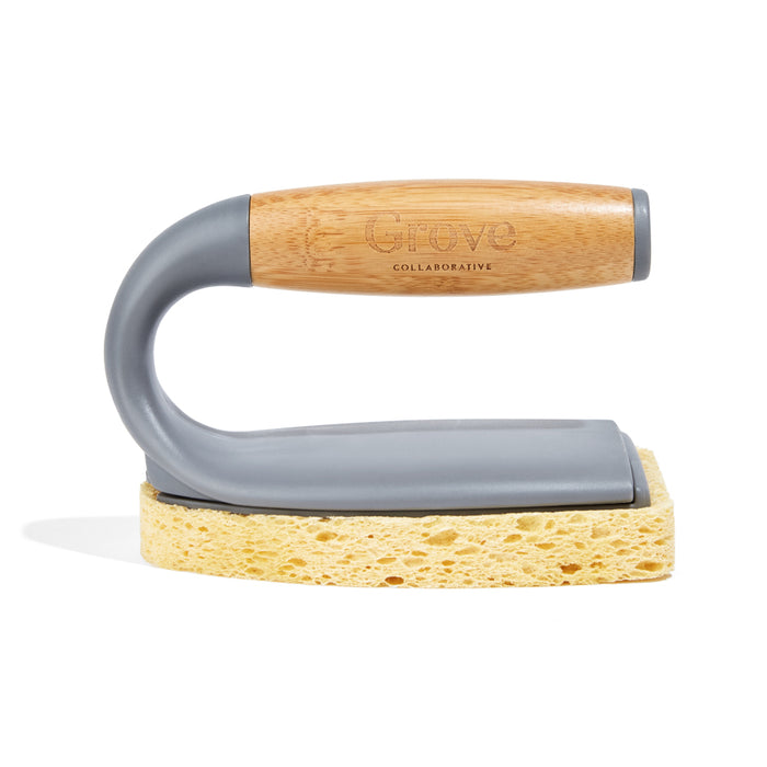 The Tub Handler + Residue Remover by Grove Co. features a scrub brush with a bamboo handle, standing upright on its gray holder and yellow sponge base.