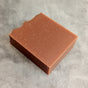 A rectangular bar of brown soap with a slightly textured surface and one wavy edge, placed on a light gray marbled background.