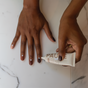 Two hands are depicted, with one applying PAUME All-in-One Cuticle & Nail Cream, enriched with plant keratin, onto the back of the other hand. The biotin-strengthened nails are painted in alternating dark brown and light beige shades on a white marble background.