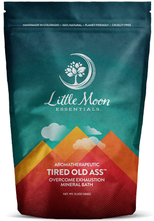 A colorful pouch labeled Little Moon Essentials Aromatherapeutic Tired Old Ass Overcome Exhaustion Mineral Bath, featuring a mountain graphic and a tree logo on a teal and orange background.