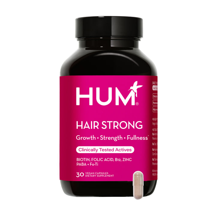 A black bottle with a bright pink label reads HUM Hair Strong. Below it says Growth, Strength, Fullness, and lists ingredients like biotin, folic acid, B12, zinc. A beige capsule is placed in front of the bottle.