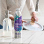 A metal bottle of Grove Co. lavender blossom and thyme dish soap stands on a kitchen counter next to a clear soap dispenser, while a person washes a dish in the blurred background.