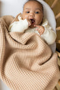 A baby in a cream-colored top lies in a crib under The Popcorn - Henna, a tan, textured, superfine merino heirloom baby blanket by AU Baby.